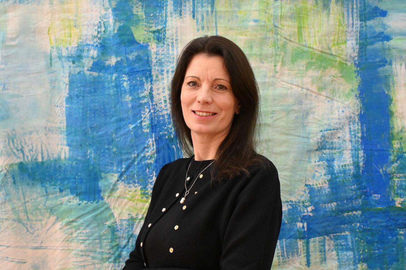 Lächelnde Frau mit schulterlangem braunem Haar und weißem Oberteil im Innenbereich.