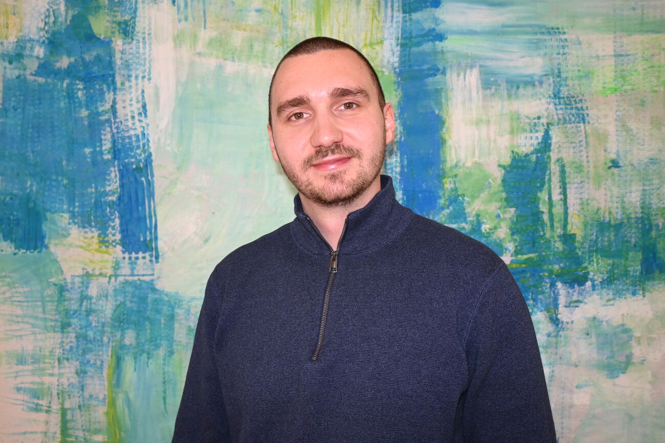 Lächelnder Mann in einem schwarzen Hemd steht vor einem blau-grünen abstrakten Hintergrund.