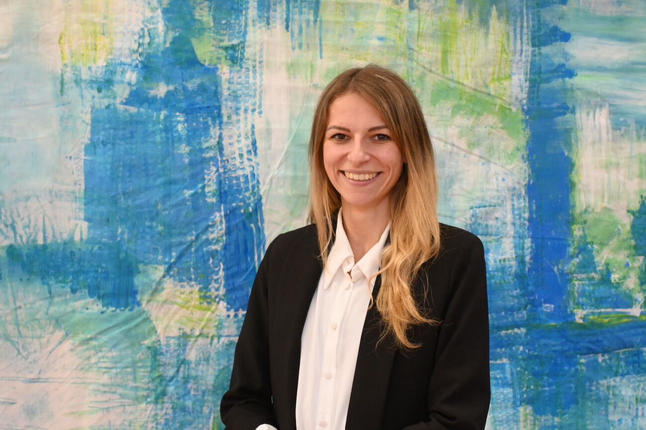 Frau mit langen blonden Haaren und dunklem Blazer posiert vor hellem Hintergrund.