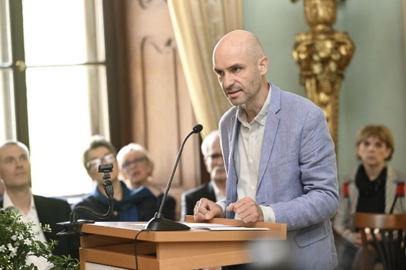 Ein Mann spricht an einem Podium vor einem Publikum in einem formellen Raum.