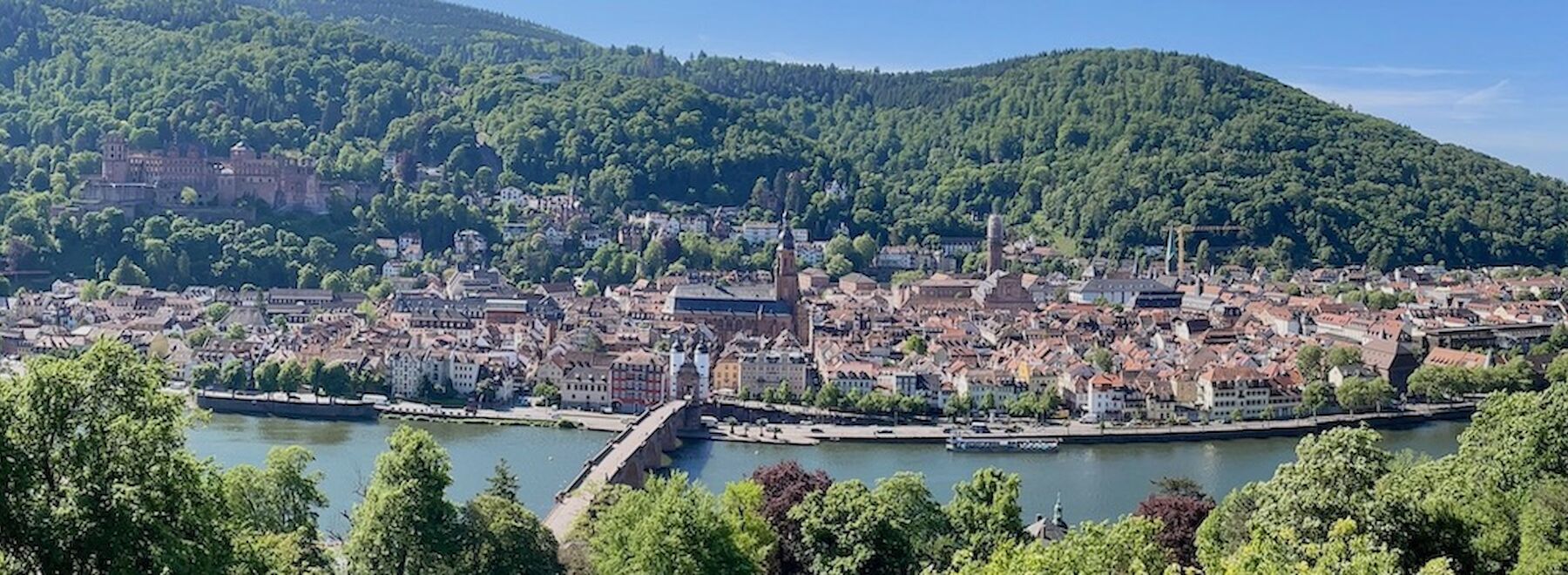 Eine malerische Stadt am Flussufer mit einer Steinbrücke und bewaldeten Hügeln im Hintergrund.