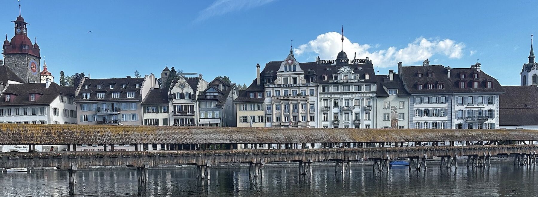 Historische Gebäude und eine überdachte Holzbrücke spiegeln sich in einem ruhigen Fluss unter blauem Himmel.