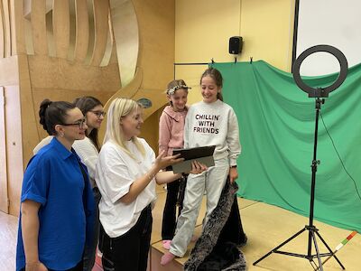 Fünf Personen lächeln vor einem Greenscreen und einem Ringlicht auf ein Tablet.