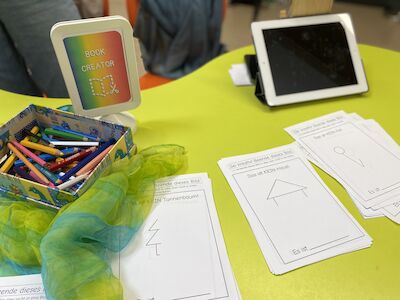 Bunte Stifte, Arbeitsblätter, ein Schal und ein Tablet auf einem leuchtend grünen Tisch.