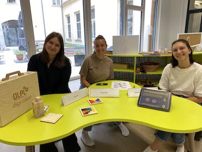 Drei lächelnde Frauen sitzen an einem leuchtend gelben Tisch, auf dem Lehrmaterialien ausgelegt sind.