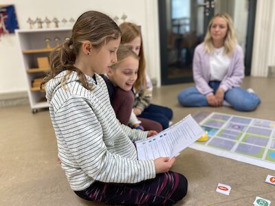 Drei Kinder lesen gemeinsam eine Zeitung, während ein Erwachsener in der Nähe auf dem Boden sitzt.