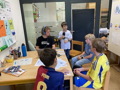 Fünf Jungen sitzen in einem Klassenzimmer an einem mit Papieren bedeckten Tisch und unterhalten sich.